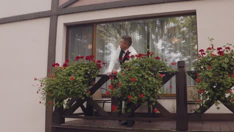 a man in a white tuxedo and a black bow tie stands on a balcony and looks away from the camera, smiling.