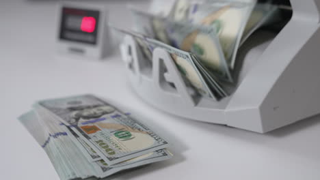 lots of hundred dollar bills are calculated in the money machine. a wad of american dollars lies in front of equipment. close up. blurred backdrop.