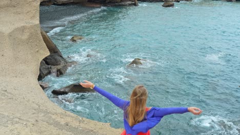 Wide-shot-of-woman-raising-arms-facing-ocean-waves,-windy-day,-tilt-up,-day