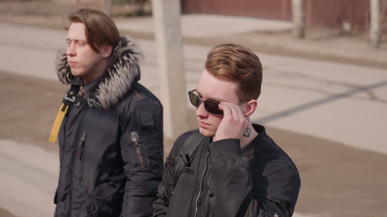 Two young men walking outdoors on city street during cold weather, one in winter jacket with fur hood looking ahead, other in sunglasses with neck tattoo adjusting glasses, casual urban lifestyle