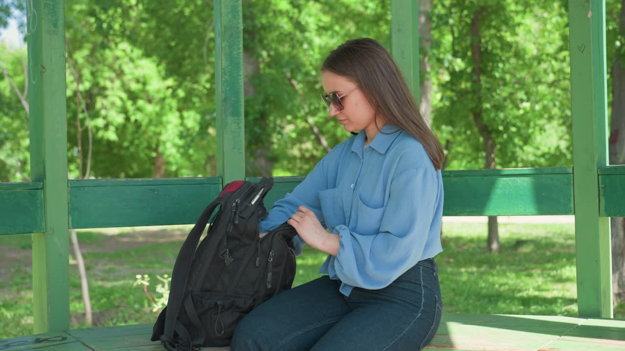 Preparativos para estudiar al aire libre: persona revisando el contenido de su mochila antes de estudiar en un parque; joven estudiante preparando material educativo sentado en un banco verde del parque.