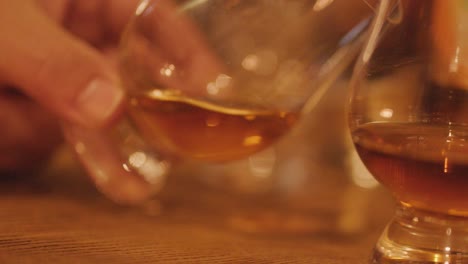 a hand gently swirls a whisky glass filled with amber spirit on a wooden table, under warm, soft lighting with shallow depth of field