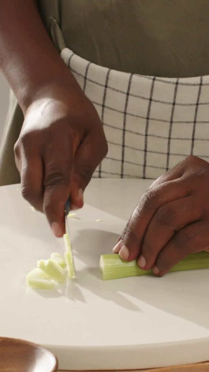 Vertical video: Placing celery, hands slicing rounds on board for meal with knife, wearing apron
