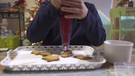 primer plano de un niño caucásico, decorando galletas de jengibre caseras navideñas con glaseado de azúcar rojo, usando una manga pastelera