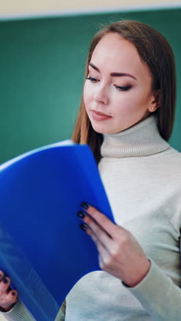 beautiful teacher with a folder. pretty young woman holding a blue folder and looking at files on the background of a board. education concept. vertical video