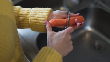 Nahaufnahmen-Einer-Frau,-Die-Einen-Gelben-Pullover-Trägt-Und-Eine-Karotte-Mit-Einem-Schäler-über-Dem-Waschbecken-Schält