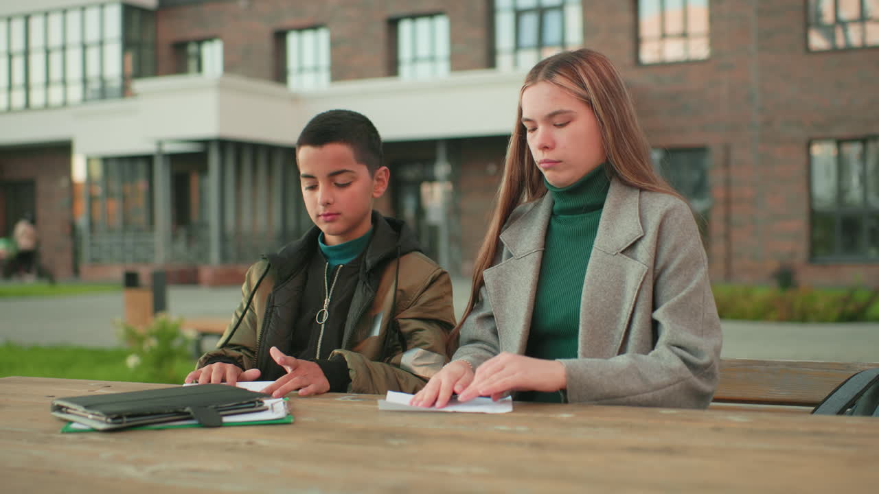 Elder sister hands brother paper while teaching him how to fold aircraft, both focused and smiling outdoors at wooden table with notebooks nearby, capturing learning, family bonding