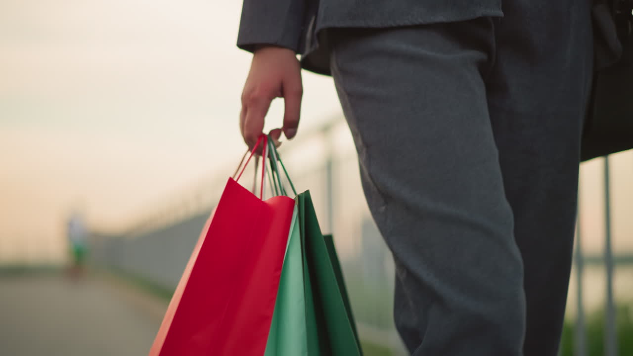 primer plano de un individuo con ropa gris que lleva tres bolsas de compras de colores mientras camina a lo largo de un carril de hierro, con un fondo borroso al amanecer