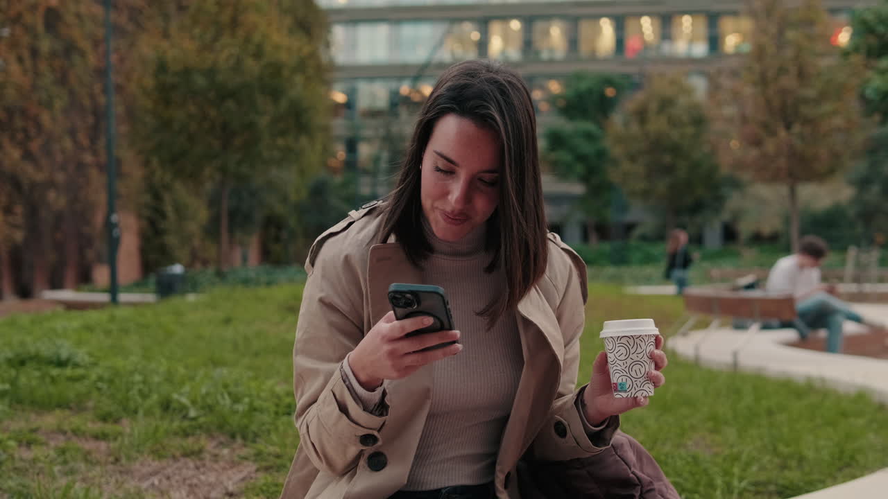 Young Woman Smiling While Using Phone and Drinking Coffee in Park