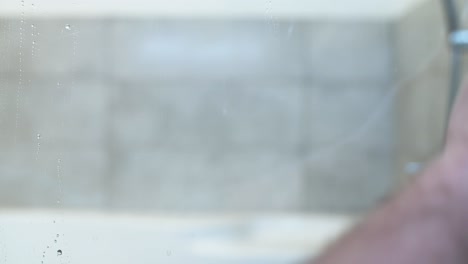 a close-up video of a person using a squeegee to clean a wet glass surface, removing water droplets and streaks. the background is blurred, suggesting an indoor bathroom or shower setting.