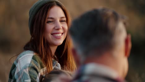 Over-the-shoulder:-A-happy-girl-in-a-green-hat-and-a-plaid-shirt-strokes-her-husband,-a-middle-aged-man-with-gray-hair,-during-her-vacation-outside-the-city-on-a-picnic-in-the-summer-evening