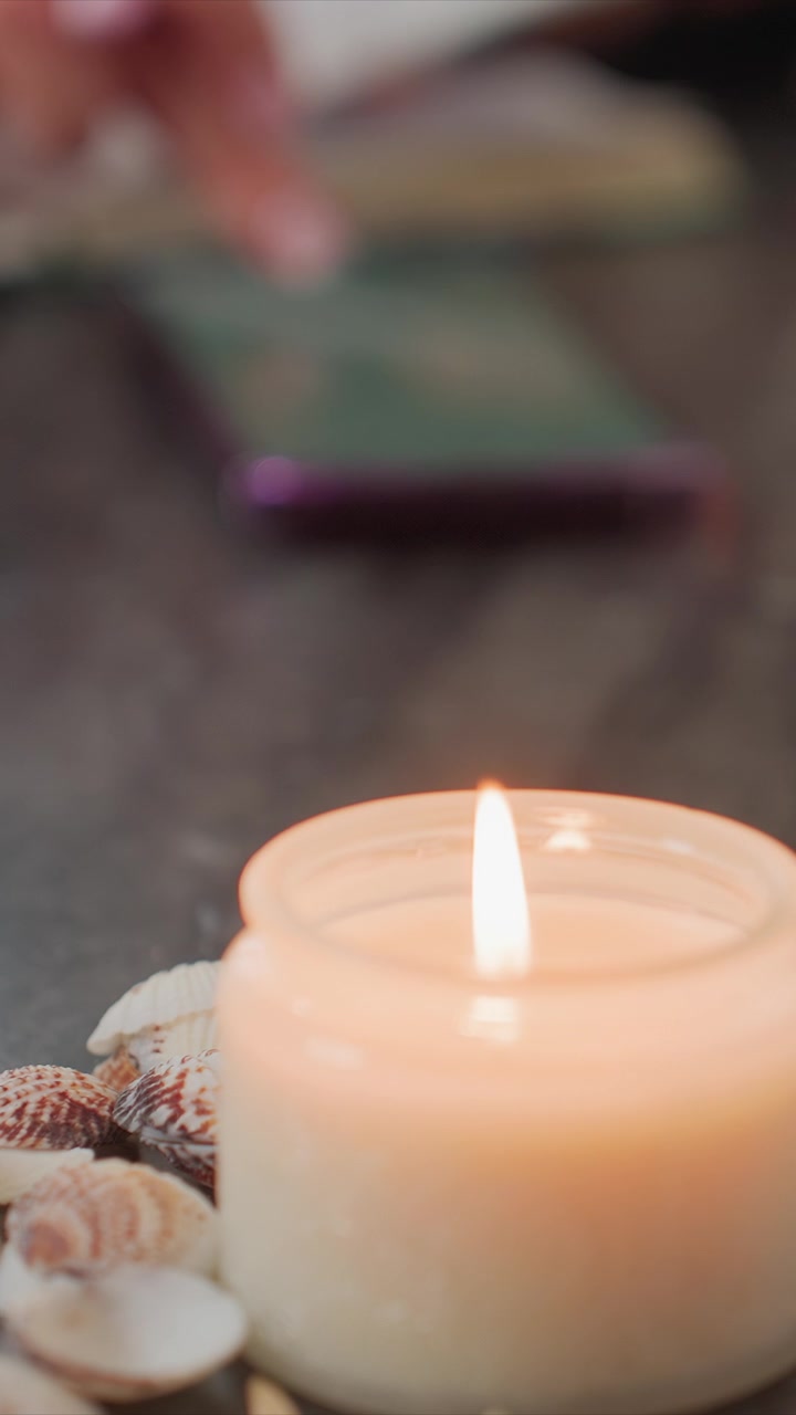 Primo piano di una candela accesa con conchiglie su una superficie scura, luce calda soffusa e profondità di campo ridotta. Lo sfondo include un libro verde sfocato o un tablet, conchiglia