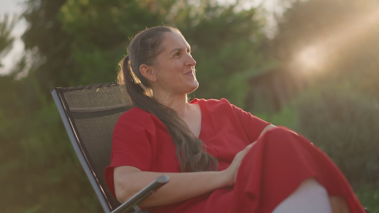 Mulher caucasiana de vestido vermelho reclinada em cadeira de jardim de vime, sorrindo sob a luz quente do sol dourado, cabelos longos trançados, professora aposentada serena desfrutando da noite ao ar livre, refletindo sobre memórias e natureza