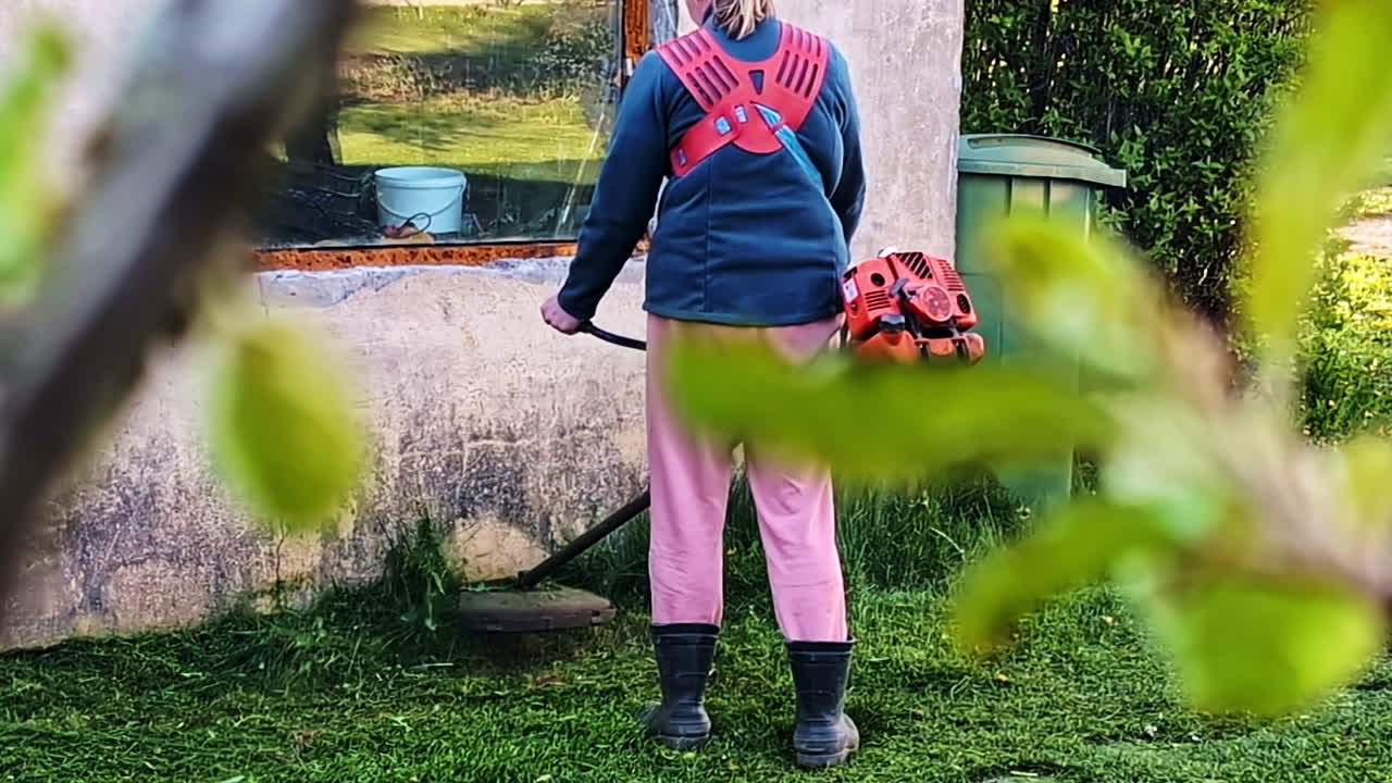 Person trimming lawn grass using brush cutter near old house with overgrown wall