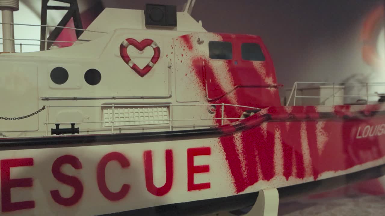 White metallic boat or ship miniature with graffiti and a heart shaped life buoy drawn on it done by Banksy on display at his exhibition behind glass under lights.