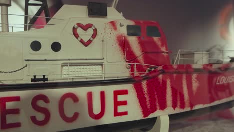 white metallic boat or ship miniature with graffiti and a heart shaped life buoy drawn on it done by banksy on display at his exhibition behind glass under lights.