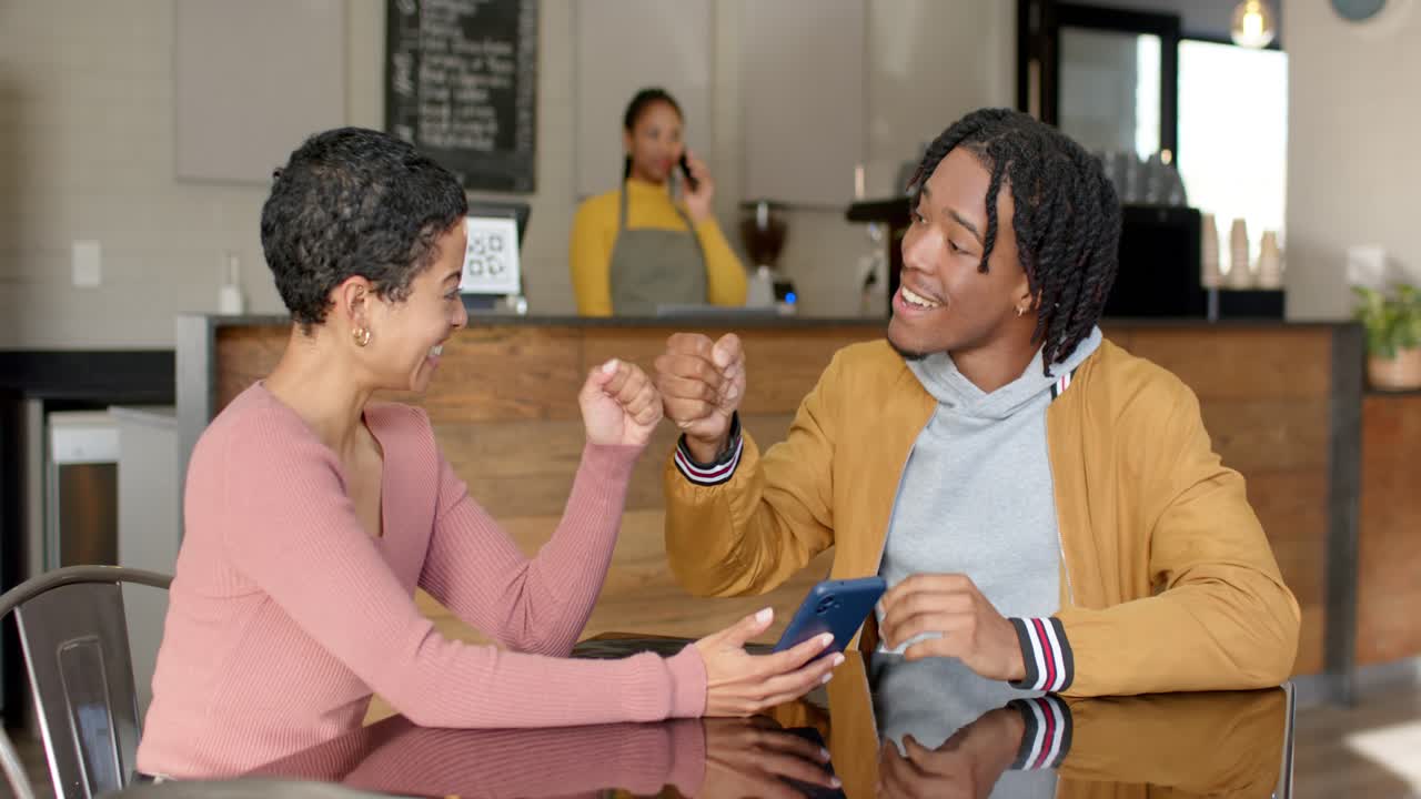 Diversi amici seduti al tavolino di un bar, una donna mostra lo schermo del suo smartphone e fa ridere un uomo che reagisce con un cenno di approvazione