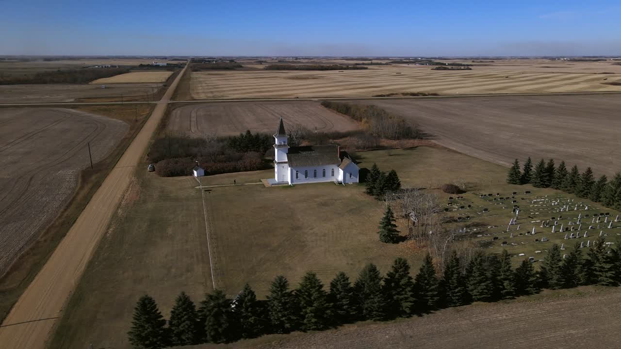 Drone orbiting around old wooden prairie chapel while person is walking along a path towards the building. St Boniface Catholic Church on a sunny day during autumn of 2021. Small cemetery adjacent