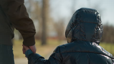 a close-up of a young boy holding hands with his father as they walk together, both dressed in warm jackets, with a blur view of trees in the background