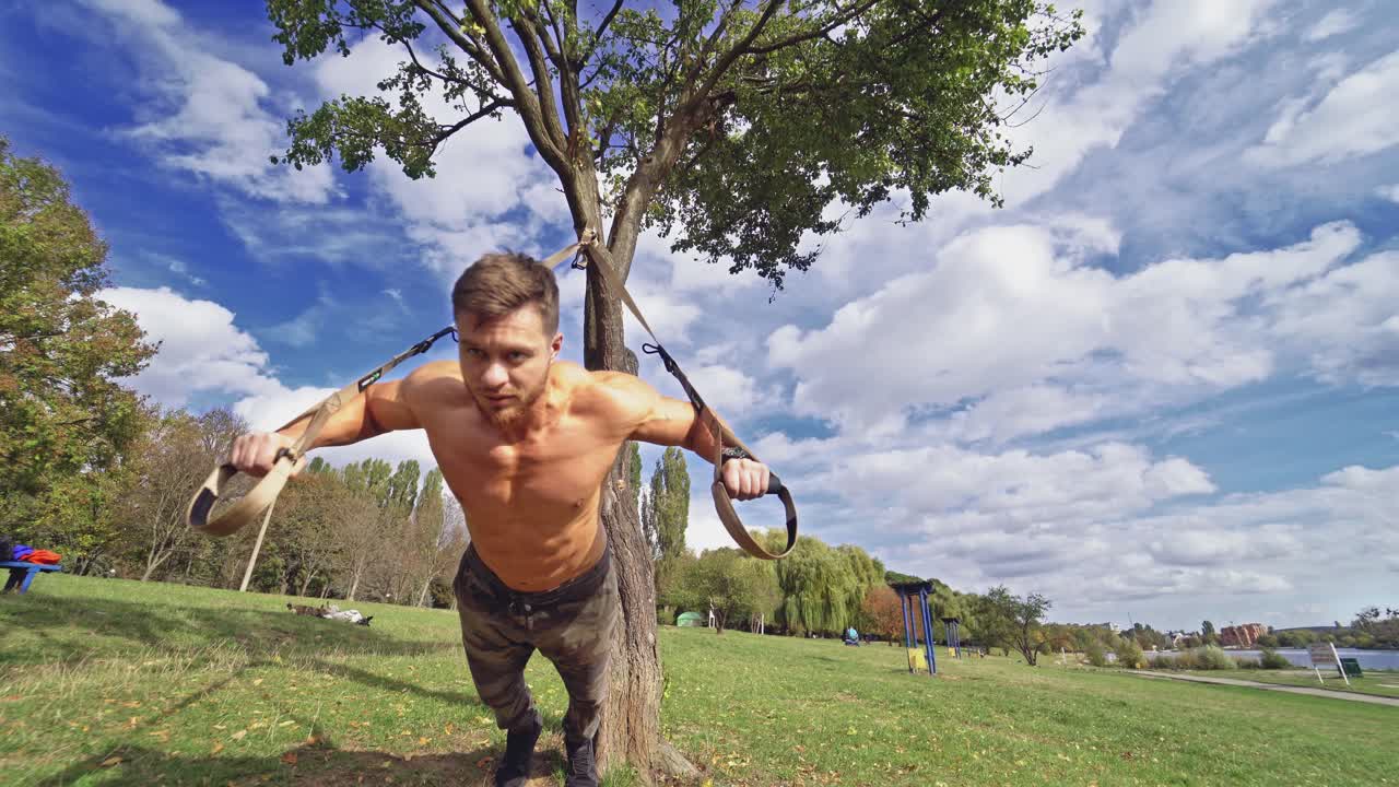 Strong bearded man having balance with trx. Topless young athlete exercising with straps outdoors in a sunny day. View from below.