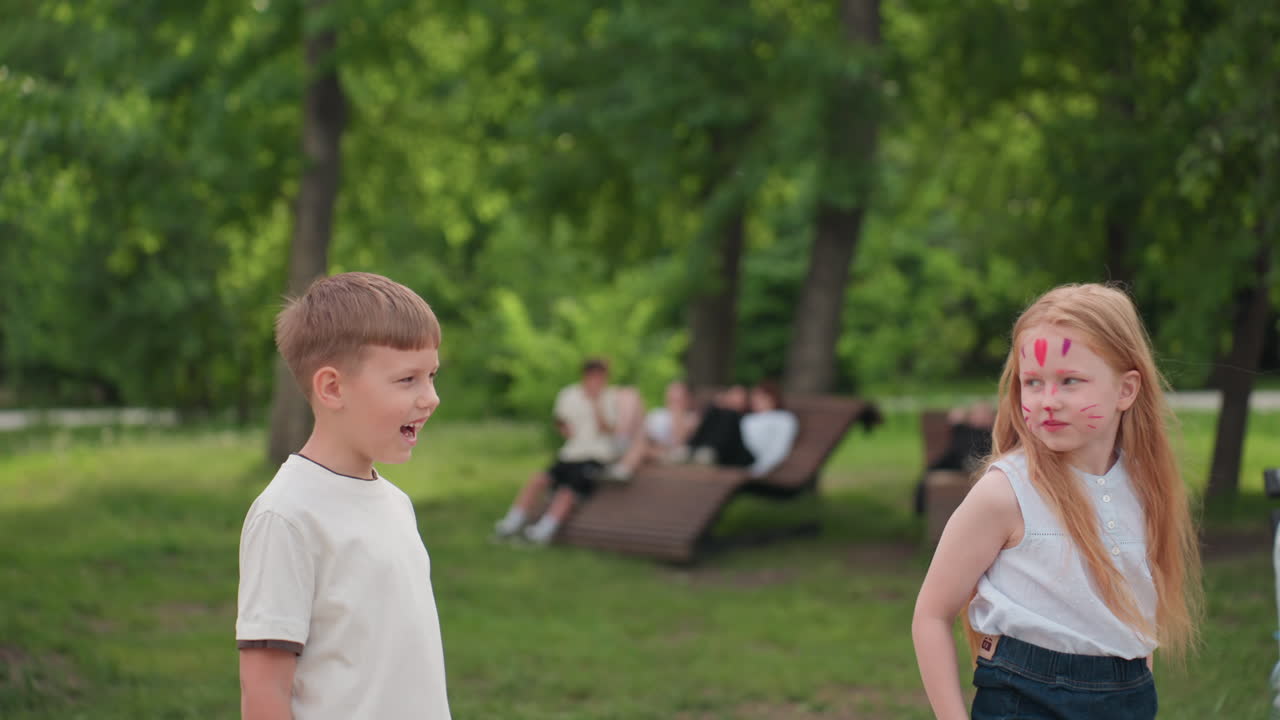 young boy and girl with face paint playing in park, blurred people sitting on bench in background, green trees framing scene, summer light casting soft glow, joyful interaction between children