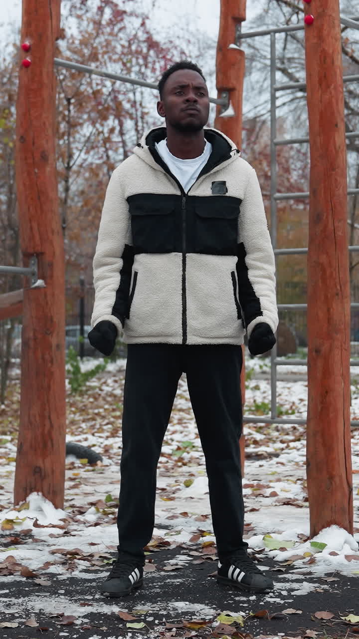 젊은 성인이 겨울 재을 입고 검은 바지를 입고 야외에서 손목 회전, 배경에 운동 장비와 함께 눈 인 공원 설정, 땅에 떨어진 잎, 멀리 지나가는 자동차