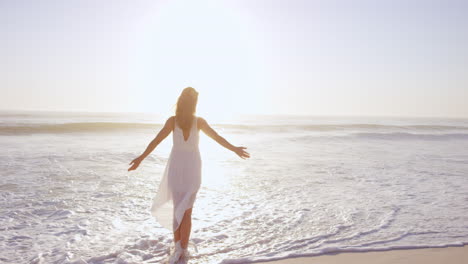 Free-happy-woman-with-arms-outstretched-enjoying-nature-walking-along-beach-at-sunset-face-raised-towards-sky