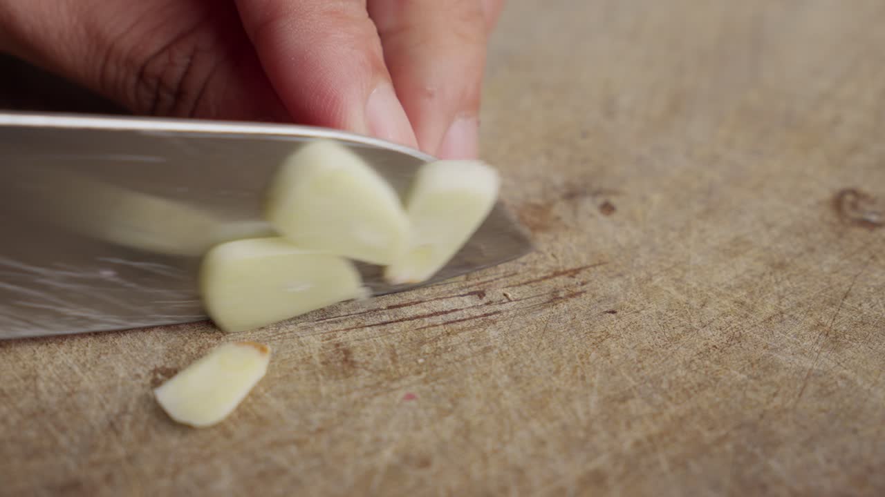 Hand reaches to hold peeled garlic and delicately chop slices on wooden cutting board, macro view