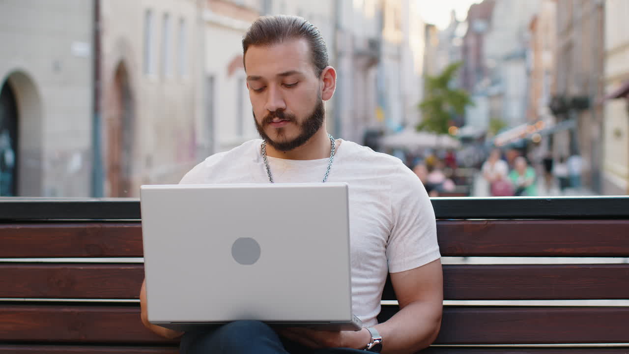Young man freelancer starts working on laptop sends messages makes online purchases on city bench