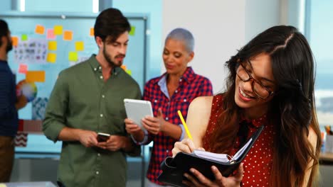 Female-executive-writing-in-organizer-and-colleagues-discussing-in-background