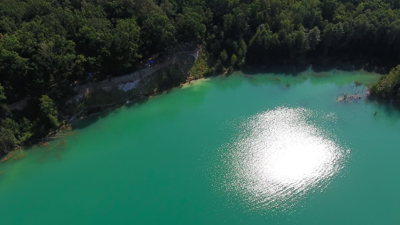 Fantastic blue water sparkling in the bright sun. Drone flying over the lake and thick woods around it. Farmlands at the backdrop.