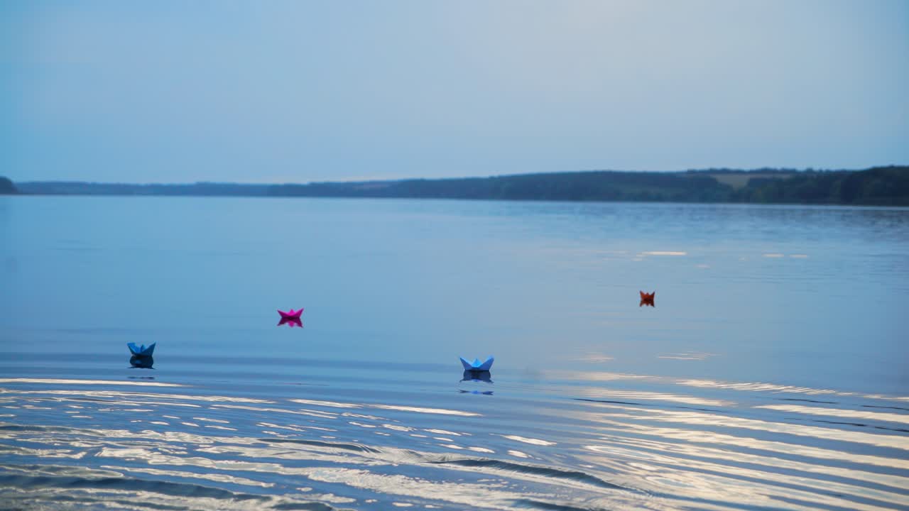 Colored paper ships go far away from the shore on the blue water background at sunset. Homemade boats with different colors floating on the big pond