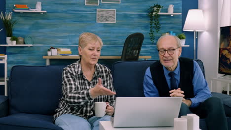 Elderly-age-couple-sitting-on-sofa-waving-at-camera-during-videocall