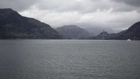Norwegian-Bridge-shot-from-a-Ferry-with-Cloudy,-Moody-weather-and-a-White-Boat-in-the-background