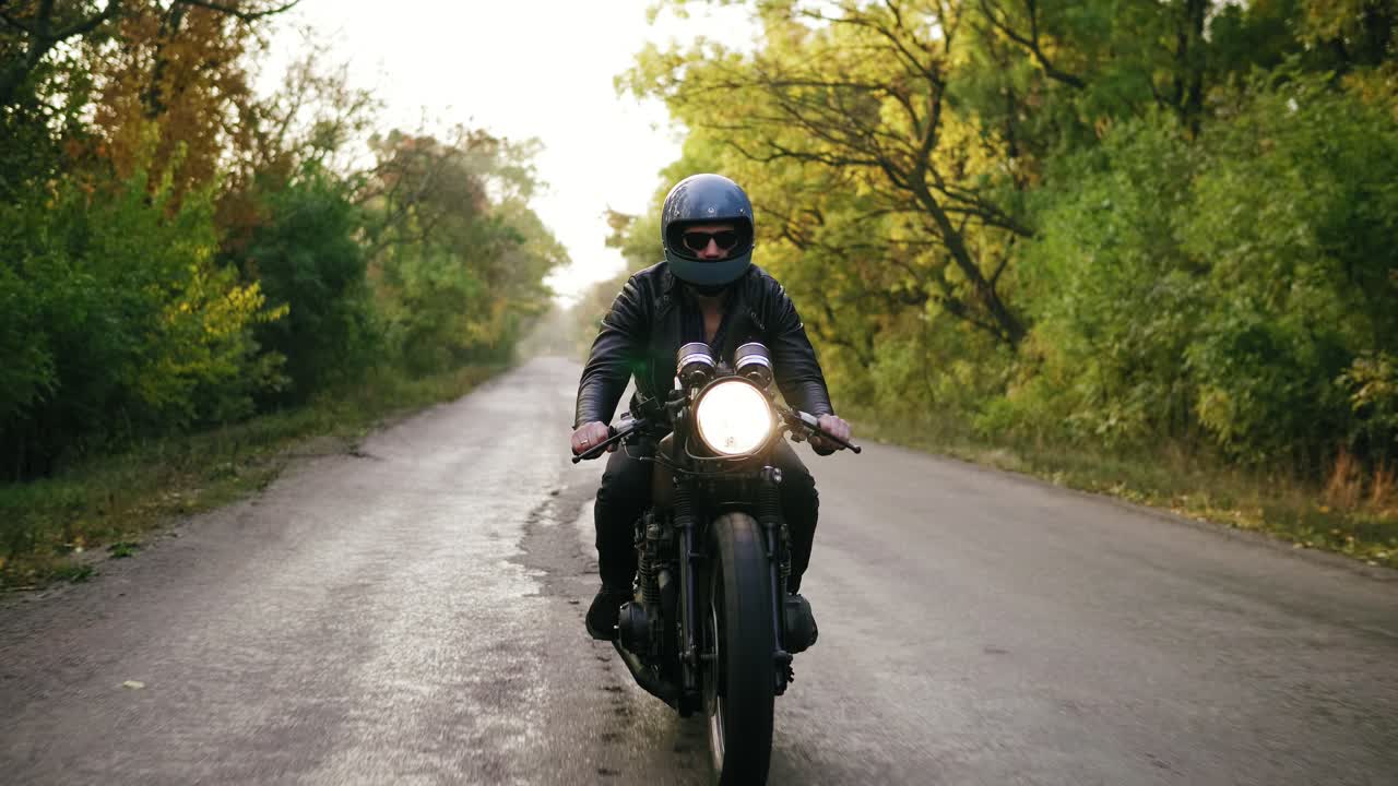 vista frontal de un joven en casco con gafas de sol y chaqueta de cuero montando una motocicleta en un camino de asfalto en otoño. árboles con hojas marrones y amarillas alrededor de la carretera