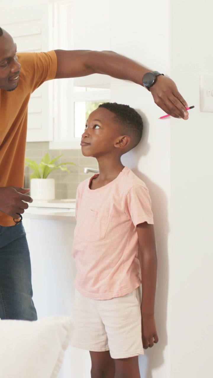 Vertical video: Measuring dad in rust tee placing hand over son at home, marking wall with pen