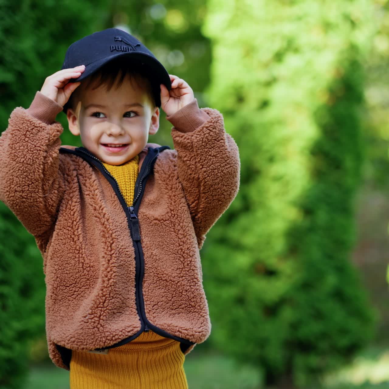 Bayi laki-laki lucu mengenakan jaket cokelat memegang topi hitam. Anak itu memakai topi menutupi dirinya. Tumbuhan hijau sebagai latar belakang
