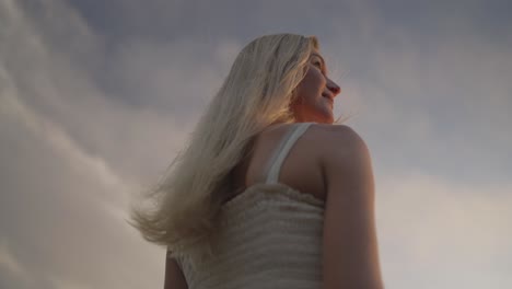 Behind-cute-smiling-blond-woman-enjoying-first-sunlight-in-morning,-clouds-in-sky,-tilt-up