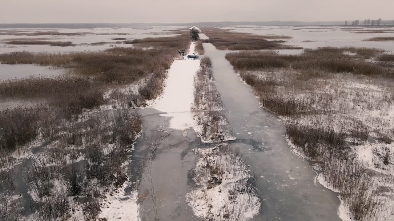 Aerial view of a frozen marshland with patches of ice, brown vegetation, and misty surroundings, creating a cold and desolate winter landscape.