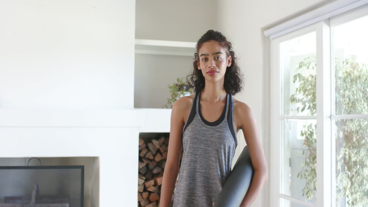 Woman entering frame from left carrying mat and dumbbell, preparing home fitness session