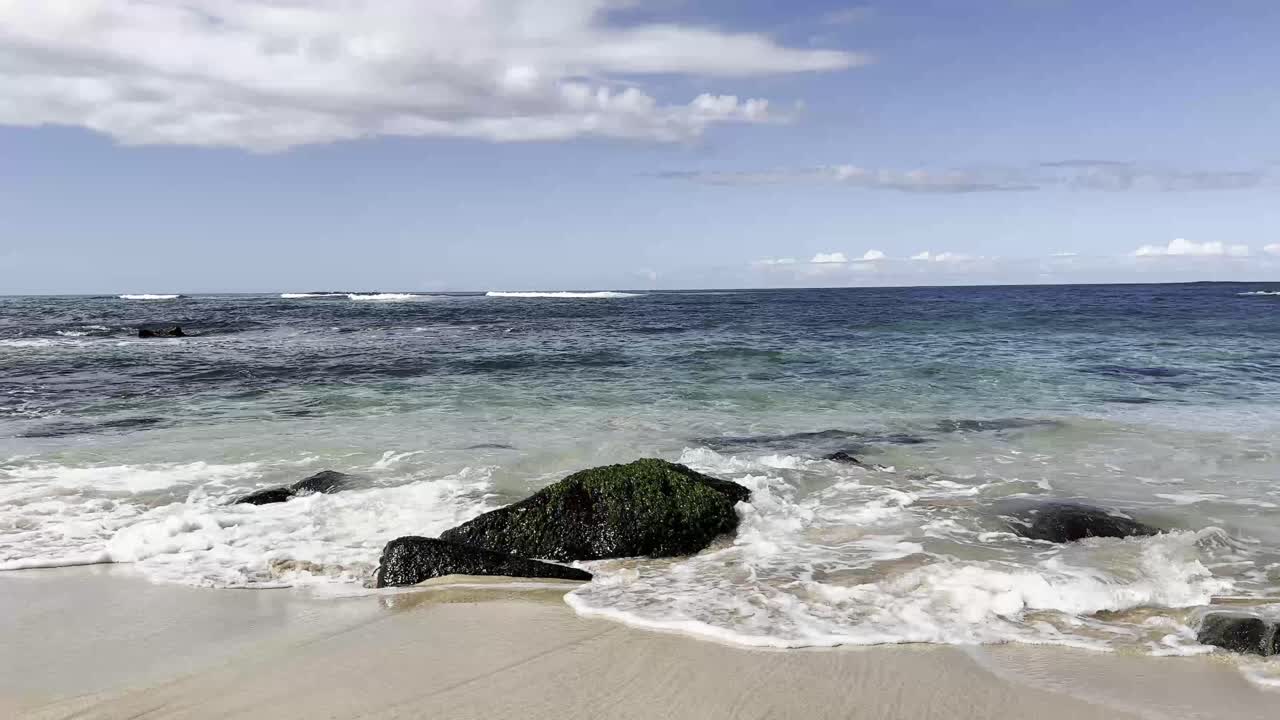 Crystal clear waves gently crash against moss-covered rocks on a pristine Hawaiian beach, capturing the serene beauty of the Pacific Ocean