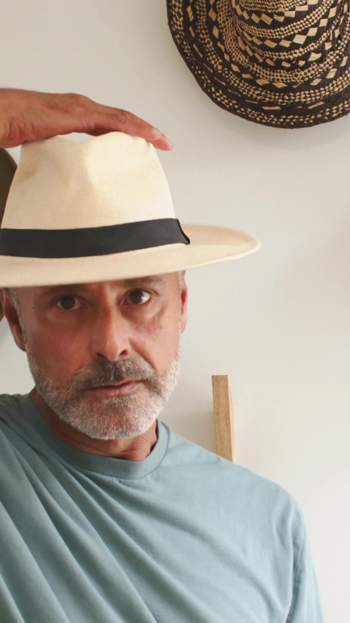 Video verticale: Uomo in maglietta blu che si allunga per provare un cappello panama, controllandone la vestibilità a casa, con cappelli appesi alla parete.