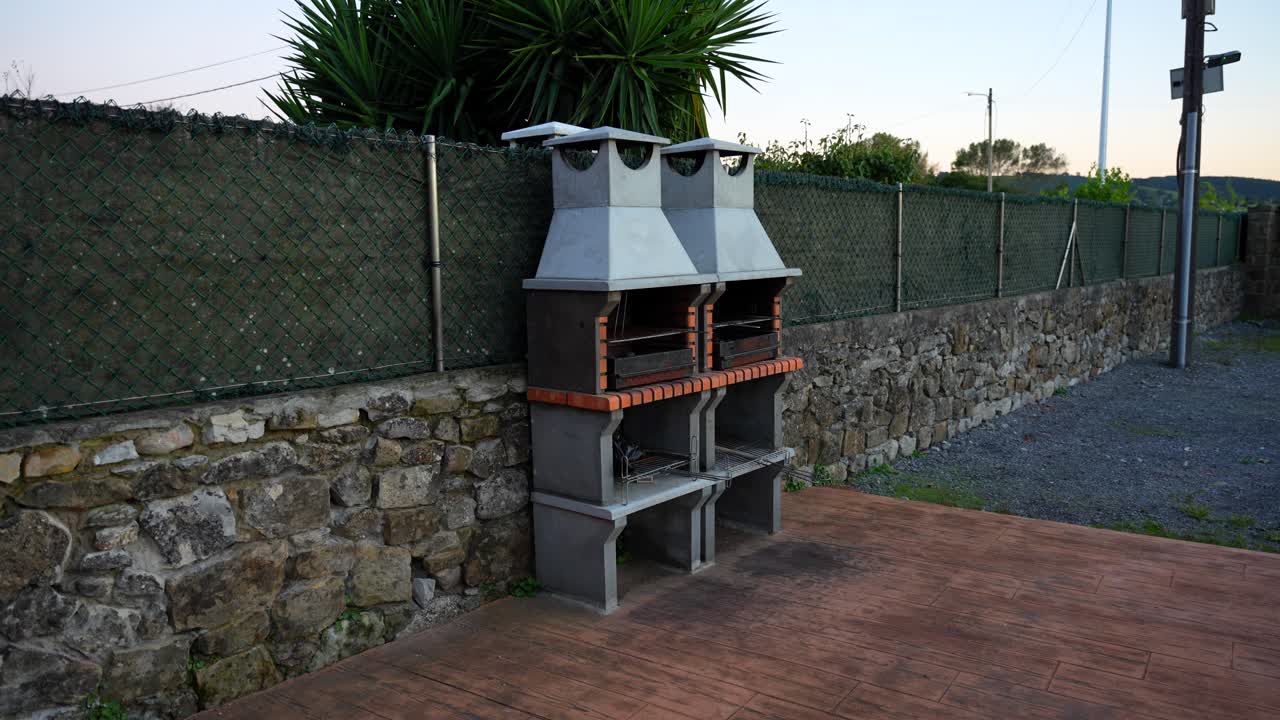 Outdoor stone grill setup against a fence in a rustic setting at dusk. Static