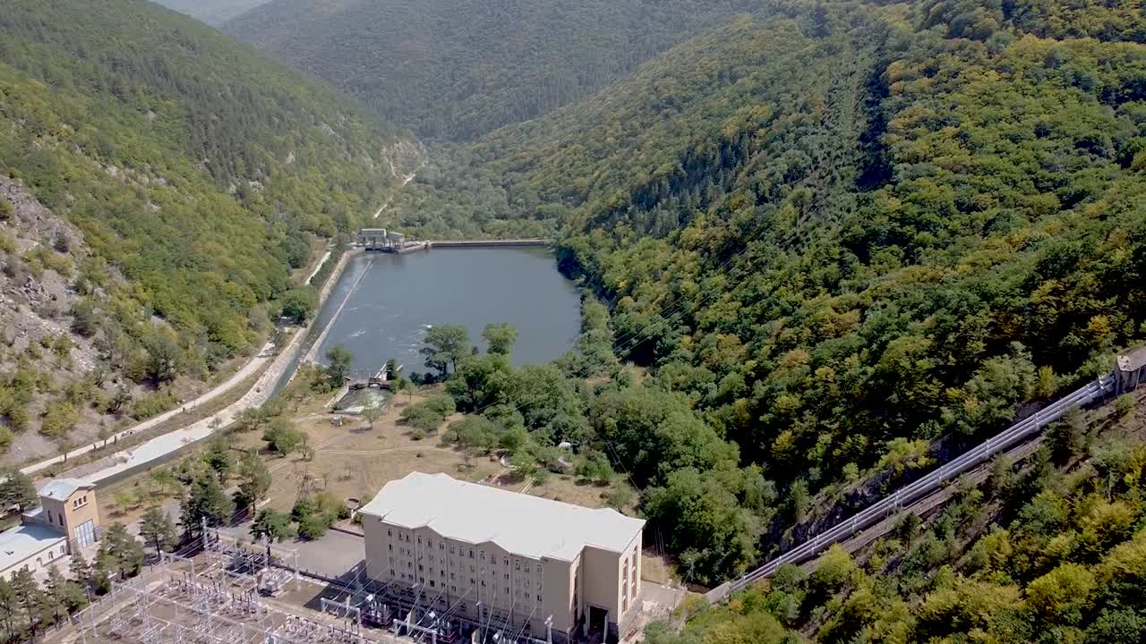A calm lake nestled in a valley, bordered by vibrant green trees and rolling hills. The sun shines brightly, casting reflections on the water, creating a tranquil atmosphere