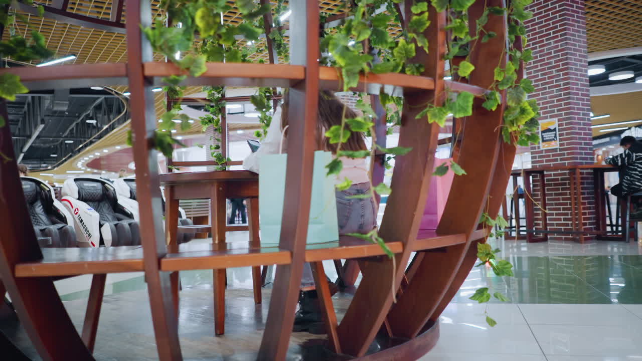 장식 된 나무 좌석에 앉아있는 젊은 여성, 에 쇼핑 가방을 들고, 다른 사람들이 쇼핑몰의 활기차고 바쁜 배경에서 고 앉아 있는 것을 볼 수 있습니다.