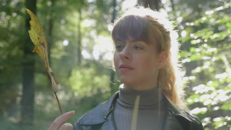 Stylish-young-woman-holds-up-leaf-in-forest-bathed-sunlight,-lens-flare