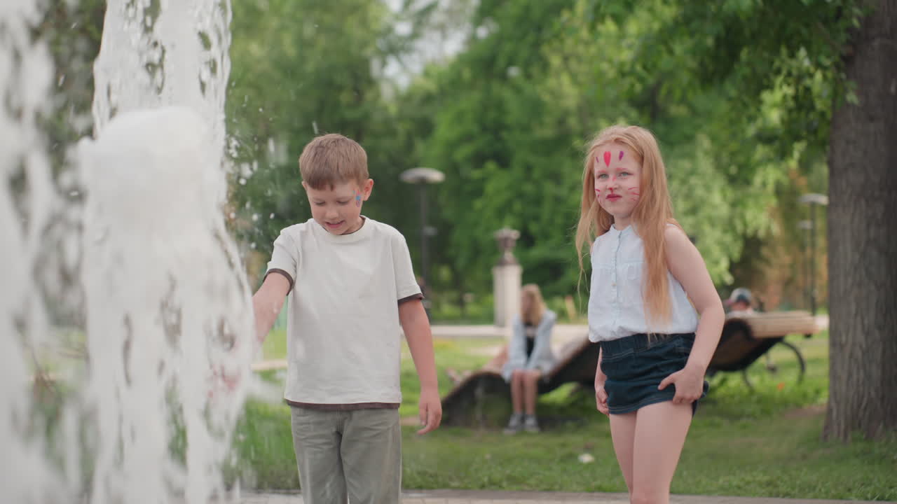 Face painted children playing with water jet in recreational park, touching splash, bench with seated people in background, summer green setting showing curiosity and joyful interaction