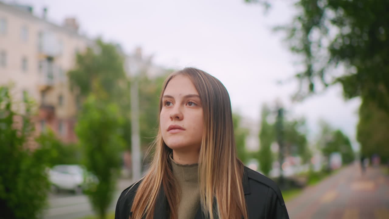 Woman with long hair in black coat looking up pensively outdoors in city park, expressing curiosity and contemplation while standing among trees with blurred urban background on cloudy autumn day