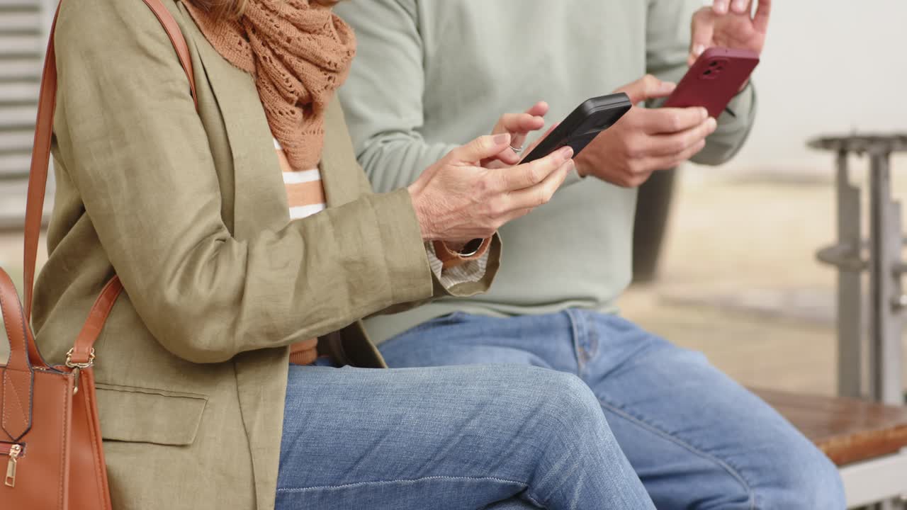 Senior couple sitting on bench in plaza tapping and swiping smartphones while checking smartwatch