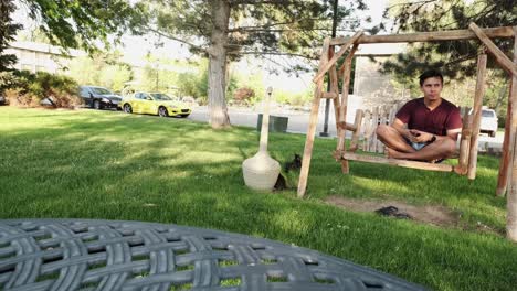 Young-Male-Person-sitting-cross-legged-on-a-hanging-bench-at-a-public-park-thinking-and-looking-afar